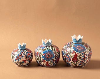 May include: Three ceramic pomegranate-shaped vessels of varying sizes, each featuring a white base decorated with intricate floral patterns in shades of blue, red, and green, topped with a light blue crown-like stem with white dots.