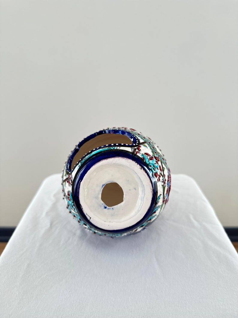 May include: A white ceramic vase with a blue and red floral pattern. The vase has a round shape and a hole in the bottom for drainage.