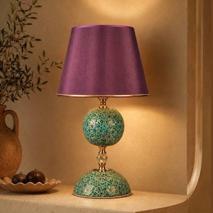 May include: A table lamp featuring a conical purple fabric lampshade and a base composed of a spherical and a semi-spherical element, both decorated with a teal, blue, and gold floral pattern, connected by a gold-toned metal stem and crystal.