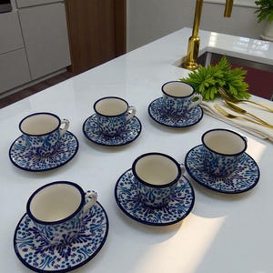 Könnte beinhalten: Sechs Keramik-Teetassen mit Untertassen, verziert mit einem blau-weißen Blumenmuster. Die Tassen haben einen weißen Innenraum und einen kleinen Henkel. Das Set ist auf einer weißen Arbeitsplatte angeordnet.