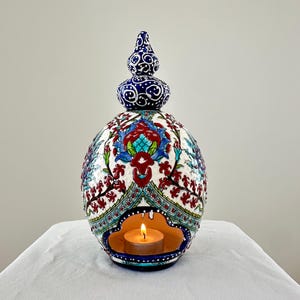May include: A white ceramic candle holder with a blue and red floral design. The holder has a small opening in the center for a tea light candle. The candle is lit and glowing.