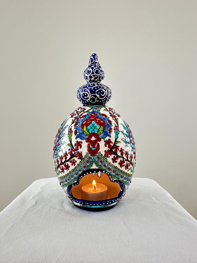May include: A white ceramic candle holder with a blue and red floral design. The holder has a small opening in the center for a tea light candle. The candle is lit and glowing.