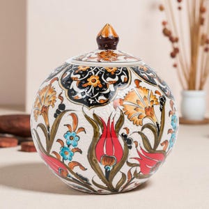 Puede incluir: Un tarro de cerámica redondo con tapa, decorado con un diseño floral. El tarro tiene una base blanca con flores rojas, naranjas y azules y hojas verdes. La tapa está adornada con un remate marrón y naranja. El tarro es probablemente para almacenamiento.