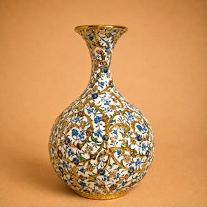 Może przedstawiać: Biały ceramiczny wazon z wąską szyjką i zaokrąglonym korpusem, ozdobiony misternym, ręcznie malowanym wzorem wijących się złotych pnączy, małych niebieskich kwiatów i zielonych liści, pokrywającym całą zewnętrzną powierzchnię.
