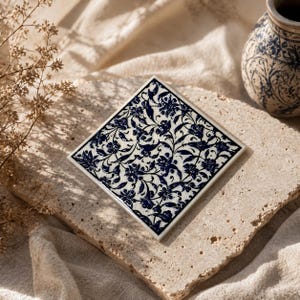 May include: A square, white ceramic tile with a navy blue floral pattern. The tile is diamond-shaped and sits on a textured, light-colored surface. A matching vase with a similar design is in the background.