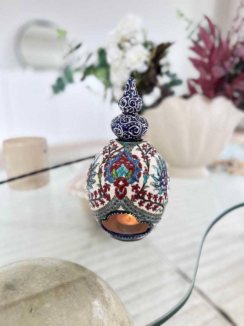 May include: A white ceramic candle holder with a blue and red floral design. The holder has a round shape with a pointed top and a small opening for a candle.