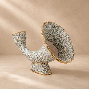 Könnte beinhalten: Eine hornförmige Keramikvase mit weißem Untergrund, einem sich wiederholenden blau-grünen Blumenmuster und einem goldfarbenen, gewellten Rand. Das Objekt steht auf einem rechteckigen Sockel und hat eine breite, trichterförmige Öffnung.