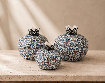 May include: Three ceramic pomegranate-shaped vases featuring a white base with intricate, colorful floral patterns in red, blue, and yellow. Each vase has a distinct black, crown-like top rim, creating a cohesive set of decorative vessels.