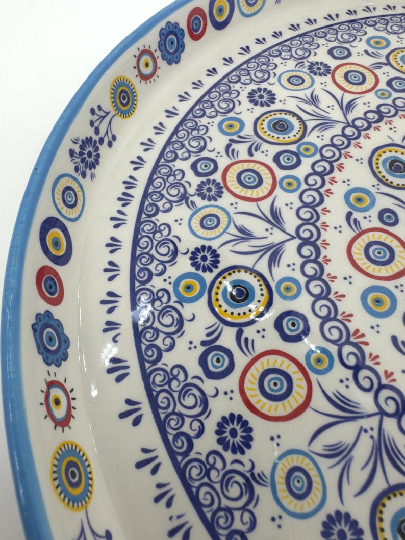 Peut inclure: Un plateau de service en c&eacute;ramique blanche avec un bord bleu et un motif d&eacute;coratif de cercles et de volutes bleus, jaunes et rouges. Le motif comprend un motif r&eacute;p&eacute;titif du symbole du mauvais &oelig;il.