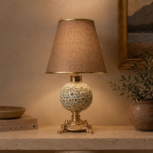 May include: A decorative table lamp with a textured brown lampshade and a gold-colored base. The lamp's spherical body features a floral pattern in blue, green, and white. The lamp is on a stone surface.