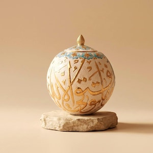 May include: A spherical ceramic vessel with a textured cream surface featuring raised gold Arabic calligraphy. The lid is topped with a gold finial and decorated with a blue and gold floral pattern, resting on a rough, light-colored stone base.
