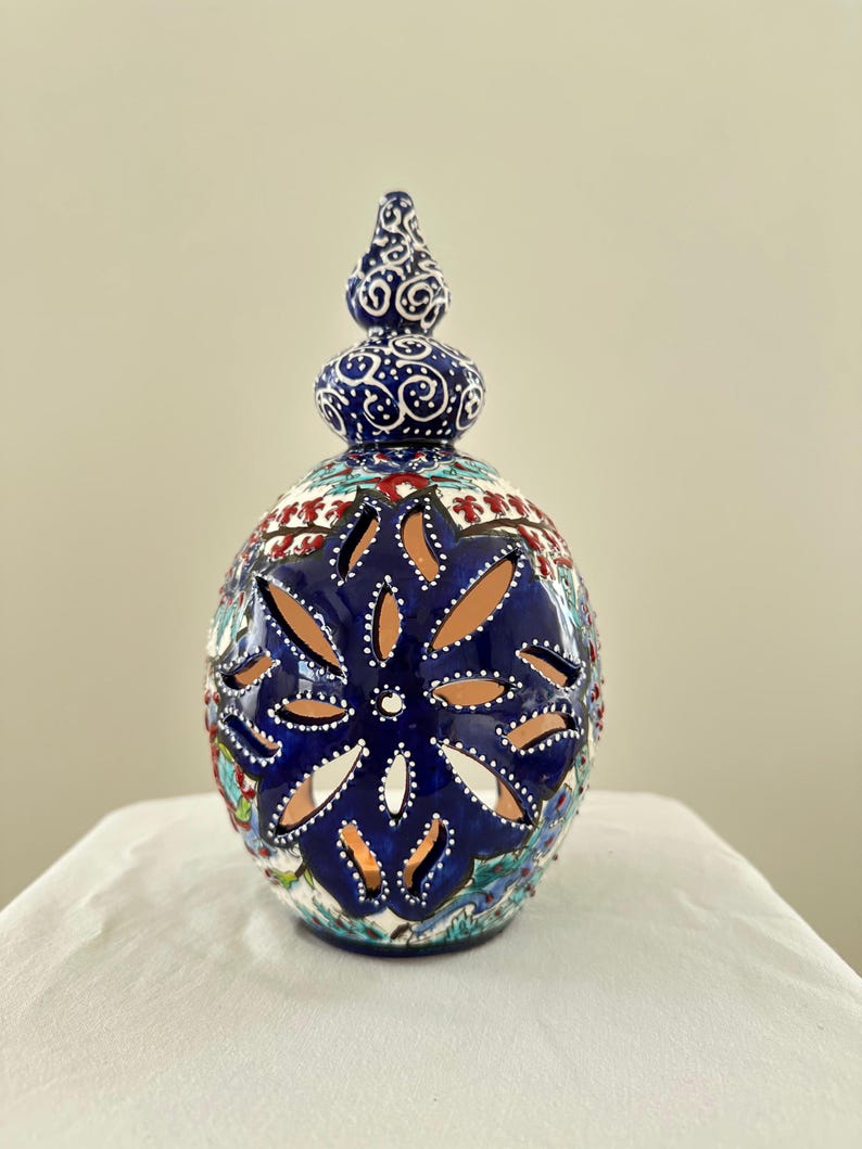 May include: A blue and white ceramic candle holder with intricate floral designs and a cut-out pattern. The holder has a domed top with a finial.