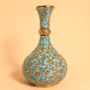 May include: A light blue vase with a narrow neck and a bulbous body, featuring an intricate, raised gold floral pattern that covers the entire surface. The design includes a textured gold band around the neck and a gold base at the bottom.