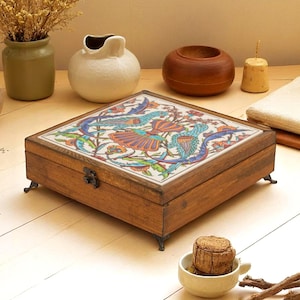 May include: A square wooden box with a hinged lid featuring a colorful tile inlay. The tile displays a detailed illustration of two birds surrounded by floral patterns in shades of blue, orange, and green. The box has a metal latch and small feet.
