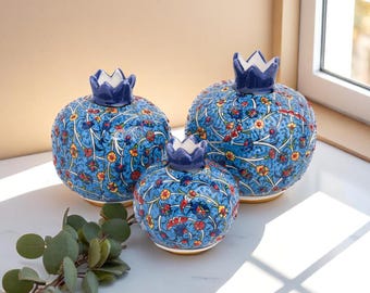 May include: Three blue ceramic pomegranate-shaped vases with floral patterns in red, yellow, and white. Each vase has a dark blue crown-shaped lid. The vases are arranged on a white surface, with a sprig of green leaves in the foreground.
