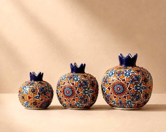 May include: Three ceramic pomegranate-shaped vessels in graduated sizes, each featuring a dark blue crown-like top and a vibrant, intricate geometric pattern in shades of orange, blue, white, and red across the rounded, textured surface.