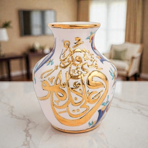 May include: White ceramic vase with gold accents and Arabic calligraphy. The vase features a flared rim and a bulbous body, with gold trim along the top and base. Decorative floral and blue accents are visible near the top. The vase is sitting on a marble surface.