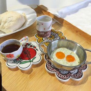 May include: A breakfast table setting with a white ceramic cup and saucer featuring a red pomegranate design, a small white ceramic cup with red hearts and a small white ceramic pan with two fried eggs on a floral patterned coaster.
