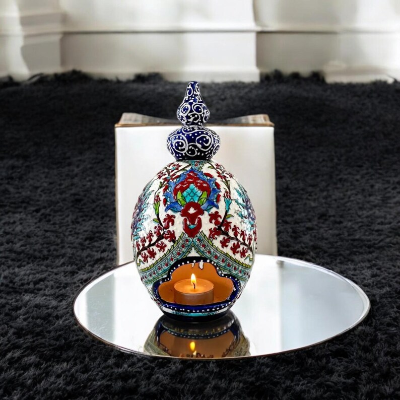 May include: A white ceramic candle holder with blue and red floral designs. The holder has a round shape with a small opening at the top for a tea light candle. The candle holder is sitting on a silver tray.