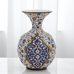 May include: A ceramic vase with a rounded body and a flared neck. The vase is decorated with a white background and detailed floral patterns in blue, orange, yellow, and red. A large blue and white medallion is centered on the front. The vase has a black base and rim.