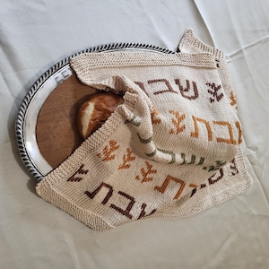 May include: A white knitted challah cover with Hebrew text and brown and green accents, covering a round loaf of bread on a silver platter.