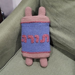 May include: A crocheted plush toy shaped like a Torah scroll with the Hebrew word "Torah" written in pink yarn on a blue background.