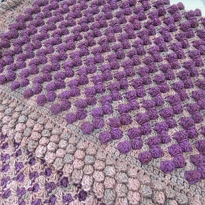 May include: A purple and brown crocheted blanket with a textured, bumpy surface. The blanket is made with a repeating pattern of squares.