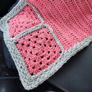 May include: A crocheted blanket with a gray border and pink squares. The blanket is made with a simple stitch pattern and has a soft, textured feel.
