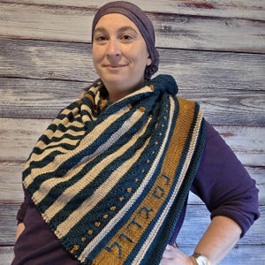 May include: A knitted shawl with a striped pattern in teal, beige, and gold. The shawl has a Hebrew inscription in gold. The person in the image is wearing a purple shirt and a purple head covering.