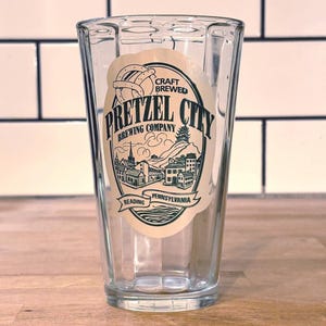 May include: A clear glass with a rectangular shape and a printed label. The label features the text "Pretzel City Brewing Company" with an illustration of a pretzel, buildings, and the words "Reading Pennsylvania". The glass is empty.