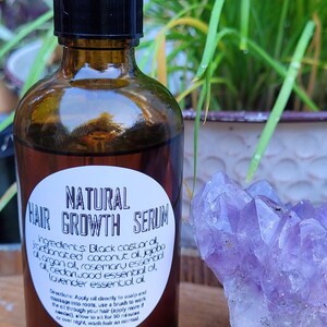 Peut inclure: Une bouteille en verre brun avec un compte-gouttes noir, étiquetée "NATURAL HAIR GROWTH SERUM". La bouteille est posée sur une surface en bois à côté d'un cristal d'améthyste violet.