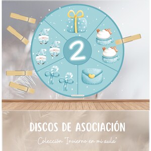 May include: A blue and white circular game board with the number 2 in the centre. The board is divided into six sections, each with a different winter-themed image: a gift box, a snowman, a candy cane, a letter, and two mugs of hot chocolate. The board is labelled "Discos de Asociación" and "Colección "Invierno en mi aula". The text "DRAGONES DE MAMÁ" is at the bottom of the board.