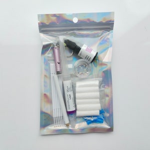 Puede incluir: Una bolsa de pl&aacute;stico transparente llena de varios art&iacute;culos para el arte de u&ntilde;as, que incluyen una botella de pegamento para u&ntilde;as morado, un peque&ntilde;o frasco de piedras de imitaci&oacute;n, bastoncillos de algod&oacute;n blancos, un bol&iacute;grafo de arte de u&ntilde;as rosa y una peque&ntilde;a botella de pegamento transparente para u&ntilde;as.