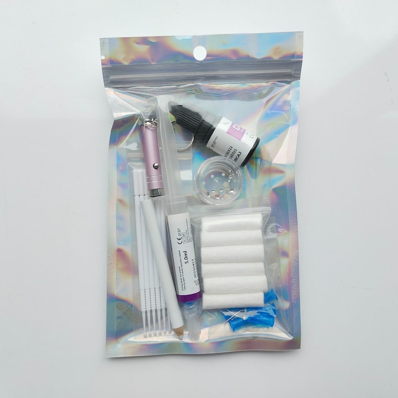 Puede incluir: Una bolsa de pl&aacute;stico transparente llena de varios art&iacute;culos para el arte de u&ntilde;as, que incluyen una botella de pegamento para u&ntilde;as morado, un peque&ntilde;o frasco de piedras de imitaci&oacute;n, bastoncillos de algod&oacute;n blancos, un bol&iacute;grafo de arte de u&ntilde;as rosa y una peque&ntilde;a botella de pegamento transparente para u&ntilde;as.