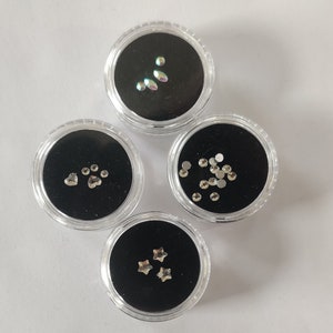 Puede incluir: Cuatro peque&ntilde;os recipientes transparentes con tapas negras, cada uno lleno de diferentes adornos plateados para u&ntilde;as. Los recipientes est&aacute;n dispuestos en c&iacute;rculo sobre una superficie blanca. Los adornos incluyen estrellas, corazones, huellas de patas y peque&ntilde;os diamantes de imitaci&oacute;n redondos.