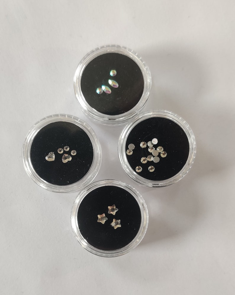 Puede incluir: Cuatro peque&ntilde;os recipientes transparentes con tapas negras, cada uno lleno de diferentes adornos plateados para u&ntilde;as. Los recipientes est&aacute;n dispuestos en c&iacute;rculo sobre una superficie blanca. Los adornos incluyen estrellas, corazones, huellas de patas y peque&ntilde;os diamantes de imitaci&oacute;n redondos.