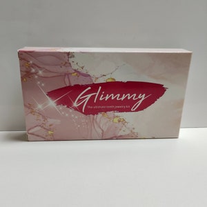 Puede incluir: Una caja rosa y blanca con un dise&ntilde;o de purpurina dorada. La caja tiene el texto "Glimmy" en letras rojas cursivas y las palabras "The ultimate teeth jewelry kit" en texto negro.