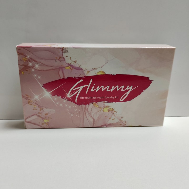 Puede incluir: Una caja rosa y blanca con un dise&ntilde;o de purpurina dorada. La caja tiene el texto "Glimmy" en letras rojas cursivas y las palabras "The ultimate teeth jewelry kit" en texto negro.