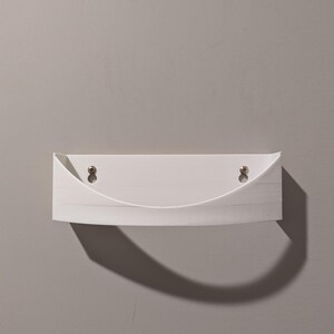 May include: White wall-mounted shelf with a curved front edge. The shelf has two small holes on the top for hanging.