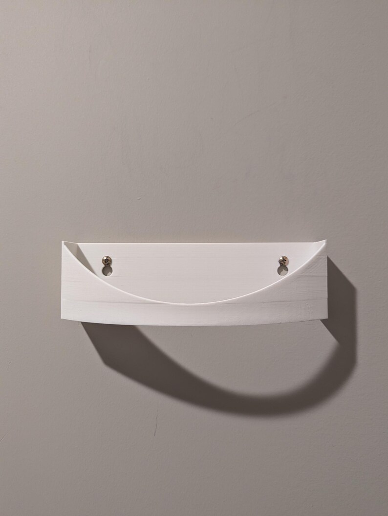 May include: White wall-mounted shelf with a curved front edge. The shelf has two small holes on the top for hanging.