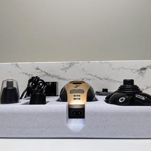 May include: A set of black and gold electric grooming tools with a charging base. The tools include a nose and ear trimmer, a beard trimmer, and a facial hair trimmer. The tools are arranged in a white foam base.