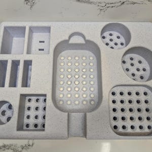 May include: A white plastic organiser with multiple compartments for storing small items. The organiser has a large, round, perforated compartment in the centre, and several smaller compartments with different shapes and sizes.