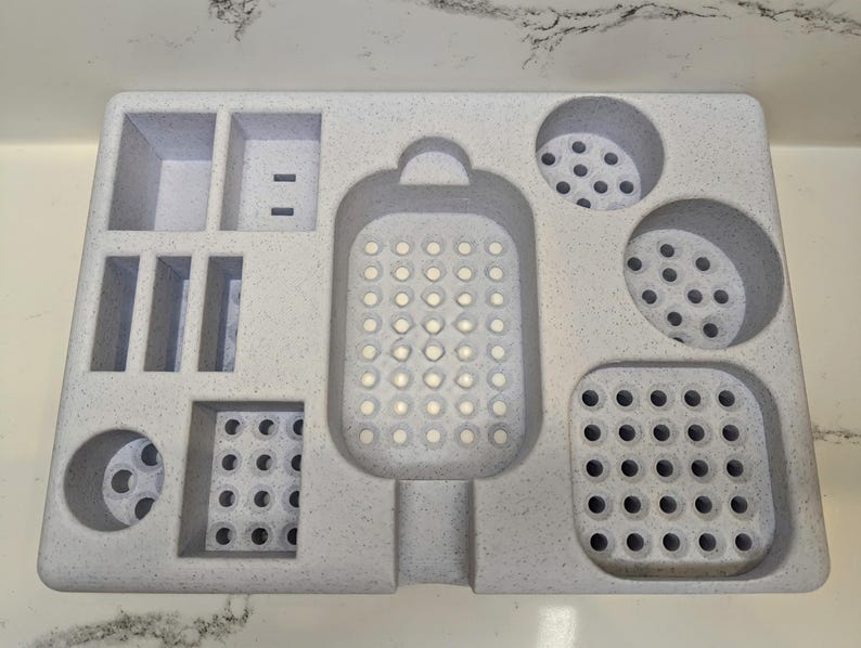 May include: A white plastic organiser with multiple compartments for storing small items. The organiser has a large, round, perforated compartment in the centre, and several smaller compartments with different shapes and sizes.