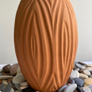 May include: A terracotta vase with a unique, textured, wavy design. The vase is sitting on a bed of smooth, gray and brown stones.