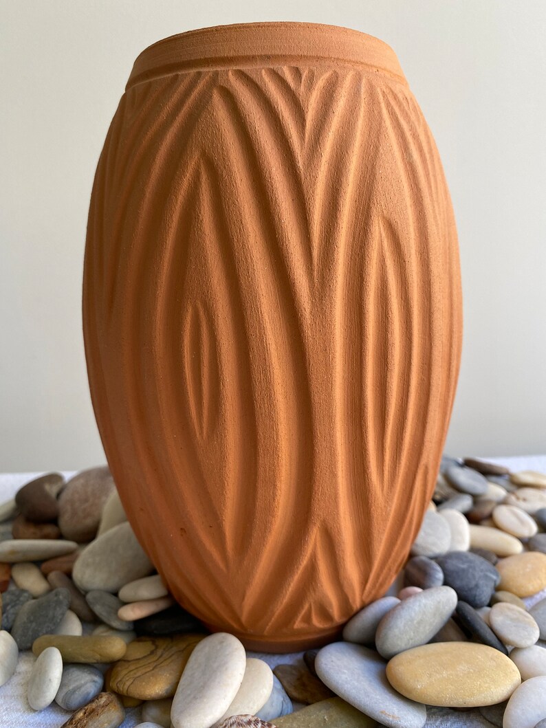May include: A terracotta vase with a unique, textured, wavy design. The vase is sitting on a bed of smooth, gray and brown stones.