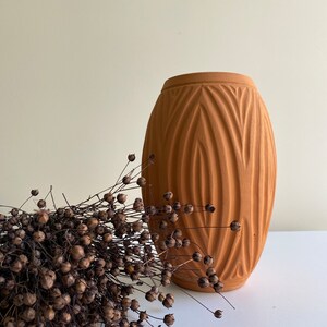 May include: A terracotta vase with a textured, geometric design. The vase is empty and is sitting on a white surface. A bunch of dried brown flax seed pods are in the foreground.