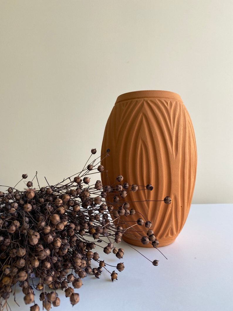 May include: A terracotta vase with a textured, geometric design. The vase is empty and is sitting on a white surface. A bunch of dried brown flax seed pods are in the foreground.