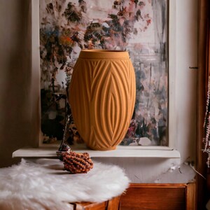 May include: A tall, orange ceramic vase with a textured, ribbed design. The vase is sitting on a white shelf in front of a framed painting with a floral design.