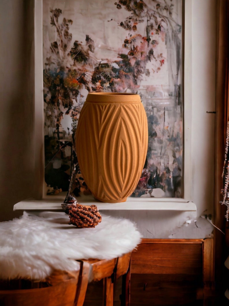May include: A tall, orange ceramic vase with a textured, ribbed design. The vase is sitting on a white shelf in front of a framed painting with a floral design.
