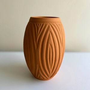 May include: A terracotta vase with a textured, geometric pattern. The vase is oval shaped and has a smooth, matte finish.